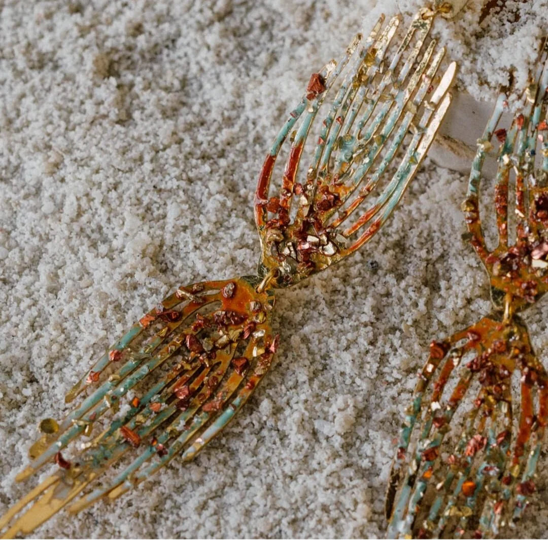 Par de pendientes artesanales de oro con forma de manos, decorados con fragmentos de cristal naranja, rojo y turquesa, colocados sobre arena