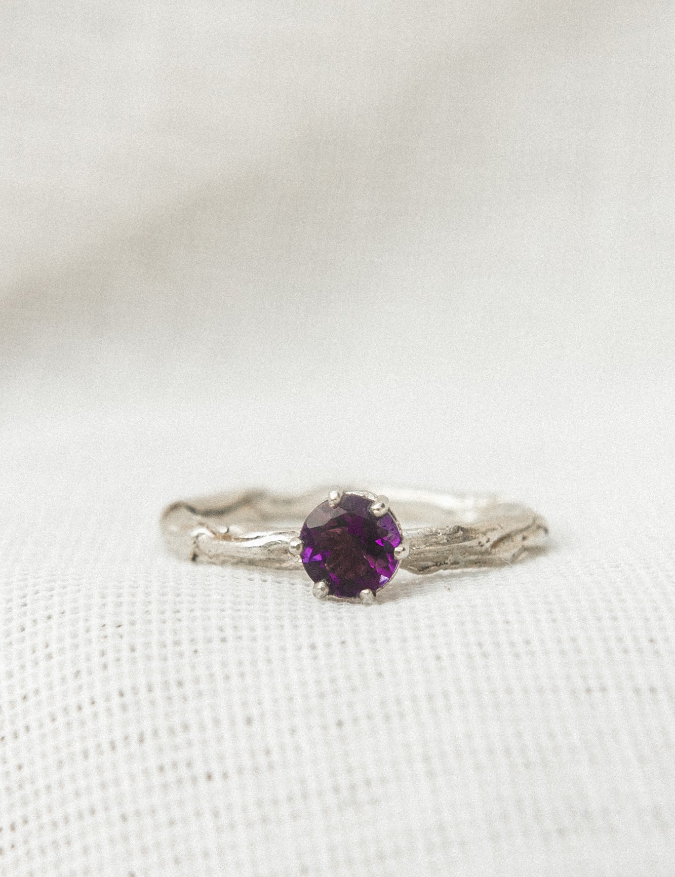 Primer plano de un anillo de plata esterlina con una amatista redonda de 4 mm montada sobre una banda texturizada, sobre tela blanca suave.