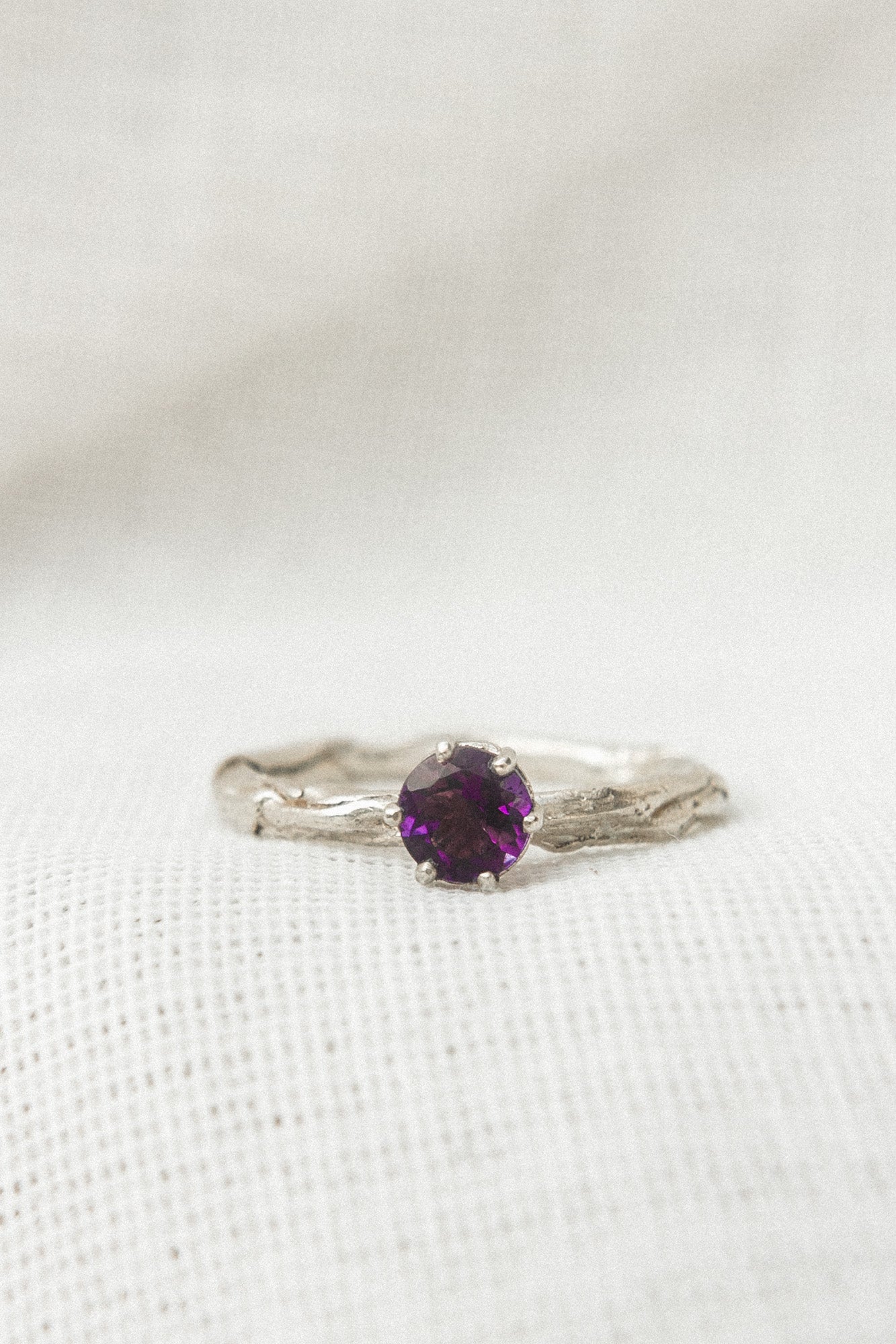 Primer plano de un anillo de plata esterlina con una amatista redonda de 4 mm montada sobre una banda texturizada, sobre tela blanca suave.