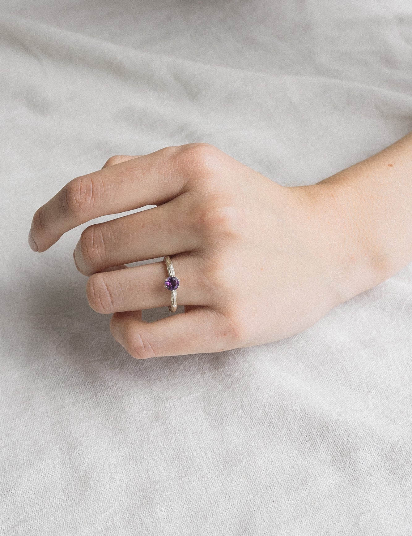 Mano de mujer luciendo el anillo de amatista en plata esterlina, mostrando el ajuste y el perfil delicado de la piedra de 4 mm sobre un fondo suave.