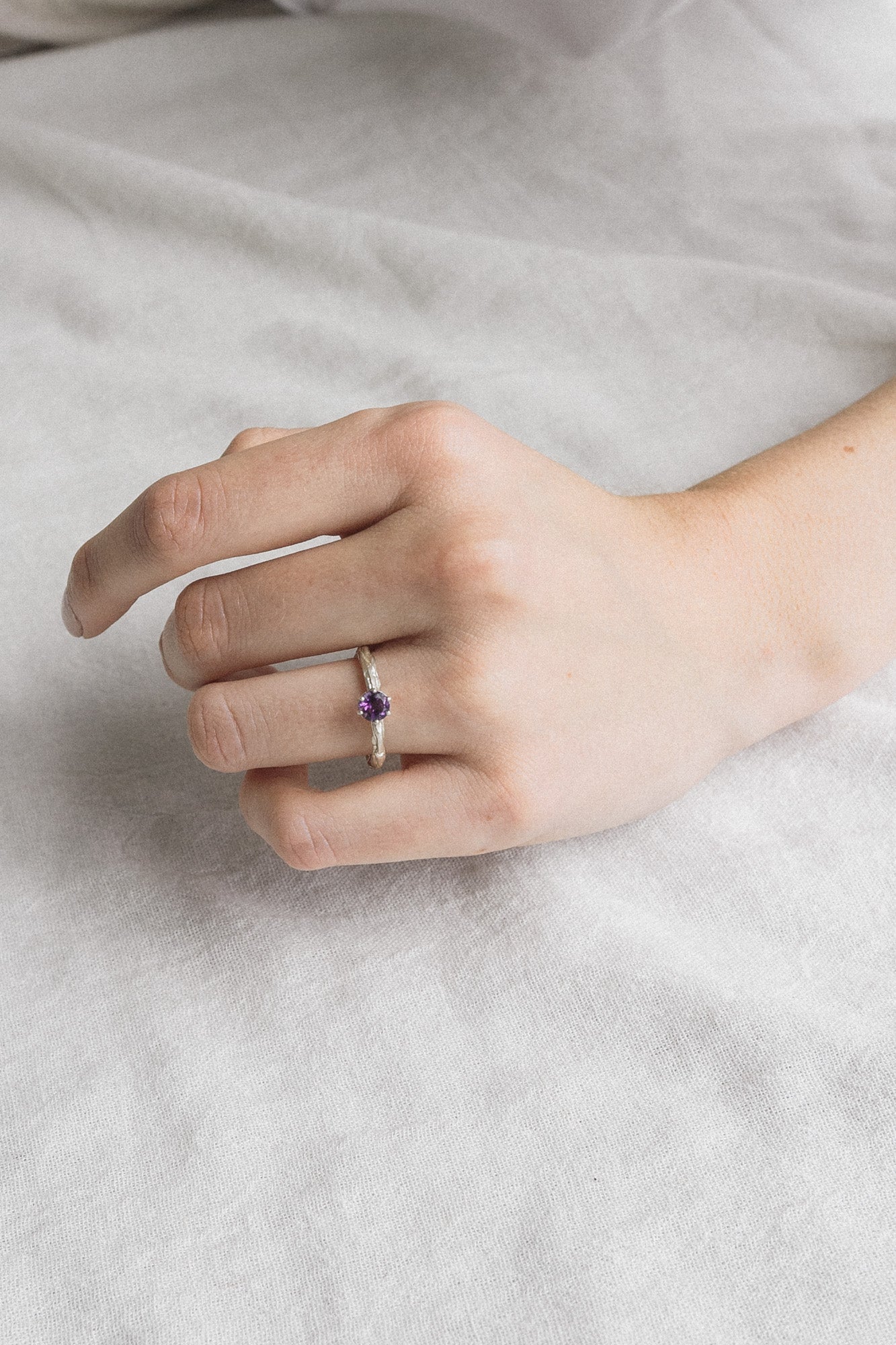 Mano de mujer luciendo el anillo de amatista en plata esterlina, mostrando el ajuste y el perfil delicado de la piedra de 4 mm sobre un fondo suave.
