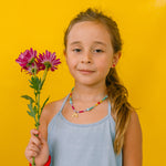 niña con collar arcoíris, sosteniendo  flores sobre fondo amarillo.