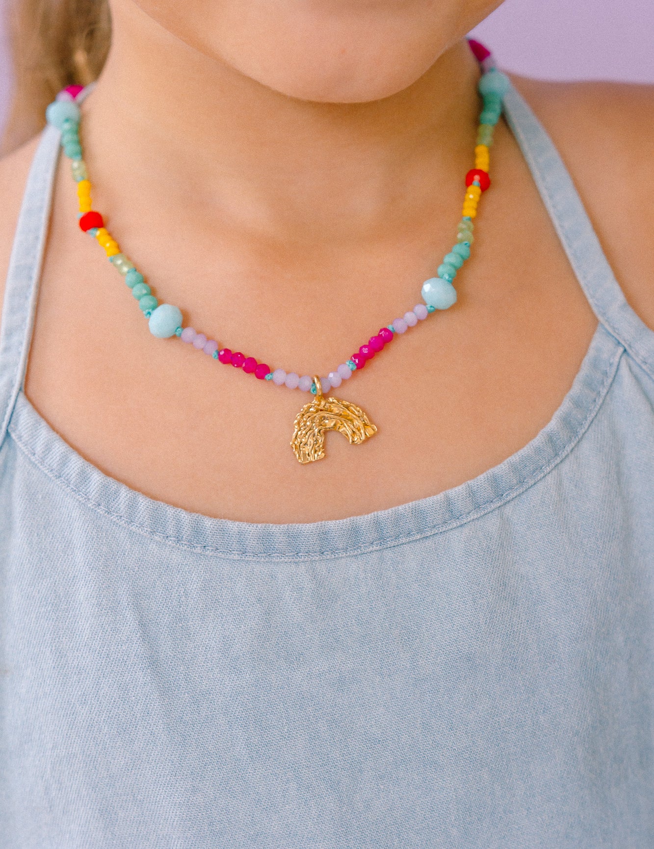Collar de cuentas multicolor con un colgante dorado en forma de arcoíris, que lleva una niña con un top azul claro.