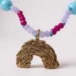 primer plano de Collar de cuentas multicolor con un colgante dorado en forma de arcoíris, sobre fondo gris claro. 