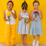 Tres niñas usando collares de cuentas multicolor, sosteniendo flores sobre un fondo amarillo.