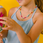 niña de medio perfil usando un collar de colores y sosteniendo una flor rosa sobre un fondo amarillo.
