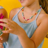 niña de medio perfil usando un collar de colores y sosteniendo una flor rosa sobre un fondo amarillo.
