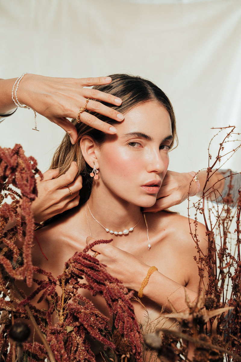 Escena estilizada con la modelo y manos ajustando los Aretes Agraz en un ambiente botánico.
