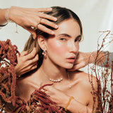 Escena estilizada con la modelo y manos ajustando los Aretes Agraz en un ambiente botánico.