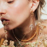 Primer plano de una modelo luciendo los Aretes Raíz, en una escena orgánica rodeada de plantas secas y tonos tierra.