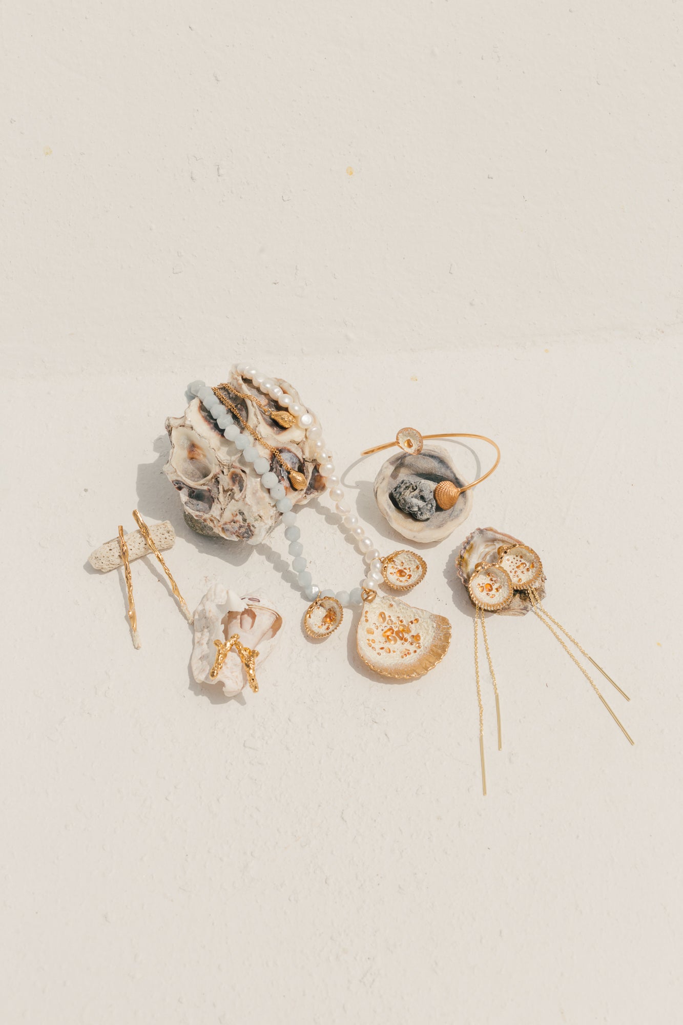 Colocación plana de joyas inspiradas en el océano, incluidas conchas bañadas en oro, collares, pendientes y horquillas de perlas y piedras preciosas, expuestas sobre una superficie de textura blanca.