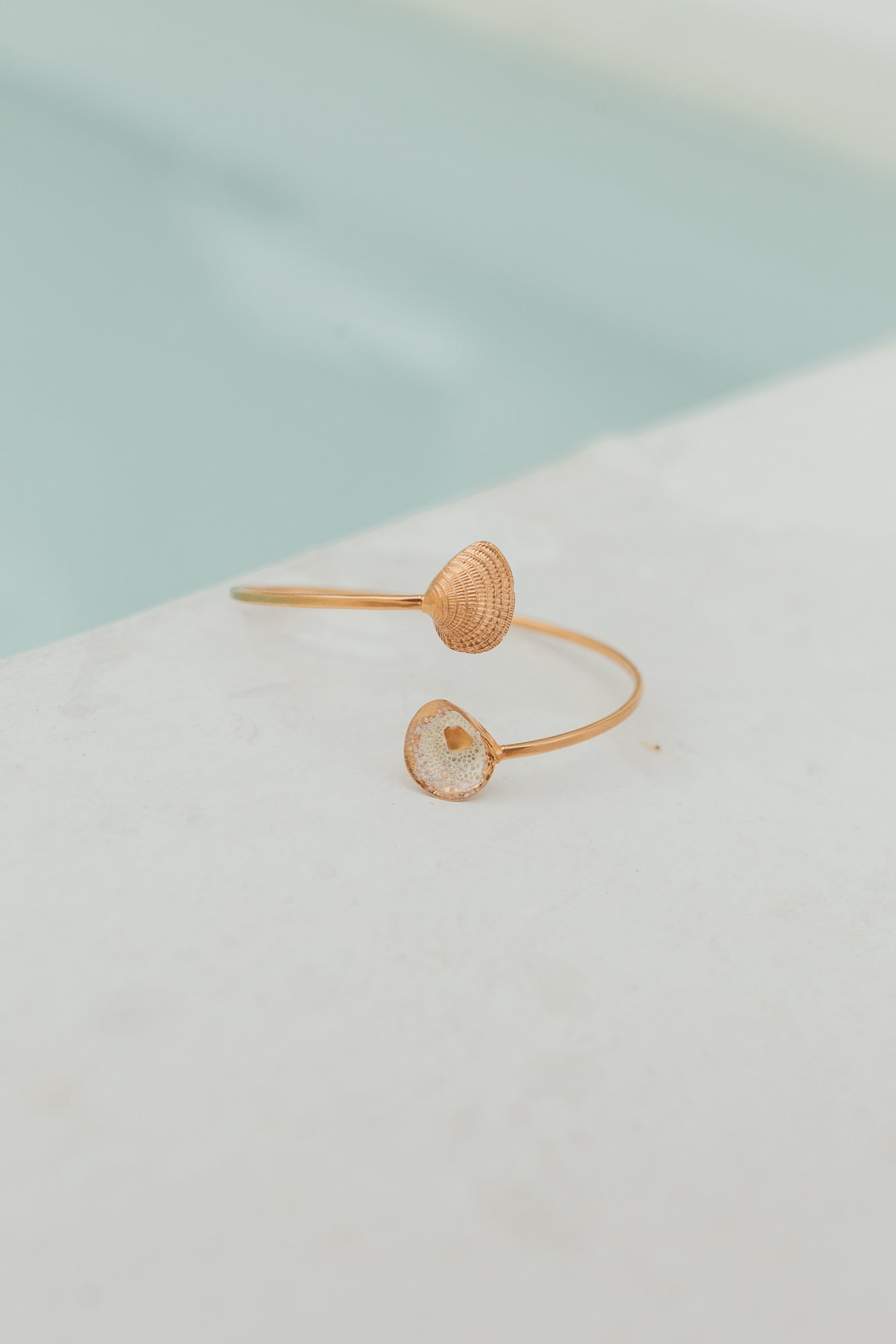 Delicada pulsera de oro con extremos abiertos que presenta una concha detallada y una concha marina rellena de resina, expuesta junto a una piscina
