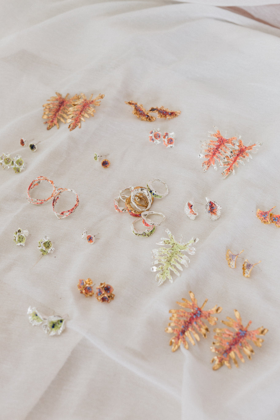 Colorido surtido de anillos y pendientes artesanales inspirados en la naturaleza, con diseños florales y coralinos en oro, plata y resinas vibrantes, expuestos sobre una suave tela blanca.