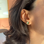 Primer plano lateral de la oreja de una mujer con un Topos Coca M en sage pintado a mano con granulación de vidrio en montura de bronce bañado en oro, cabello recogido, sobre fondo neutro.