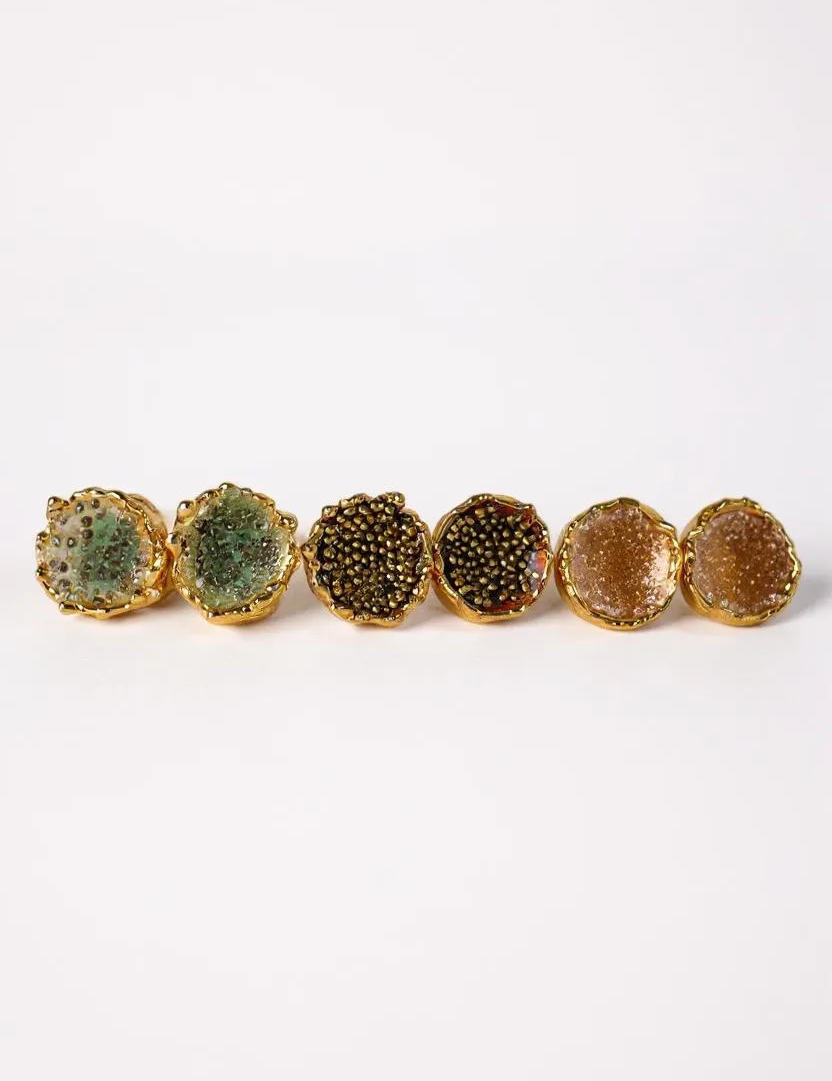 Disposición plana de seis aretes Topos Coca M sobre fondo blanco, mostrando las tres variantes pintadas a mano: dos en verde salvia con granulación de vidrio, dos en rust con granulación de vidrio y dos en tono sunset con granulación turquesa, todas con montura de bronce bañado en oro.