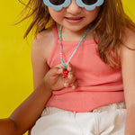 Niña con gafas de sol de montura blanca sostiene el colgante del Collar Fresa cerca de su rostro sobre un fondo amarillo.