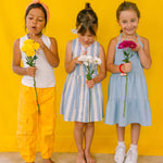Tres niñas sosteniendo flores sobre un fondo amarillo, usando callares de cuentas multicolor. 