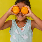 Niña con blusa sin mangas verde menta sostiene dos naranjas sobre los ojos, sonriendo frente a un fondo amarillo brillante.