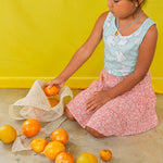 Niña arrodillada en el suelo con blusa verde menta y falda pastel juega con naranjas y lleva puesto el Collar Naranja
