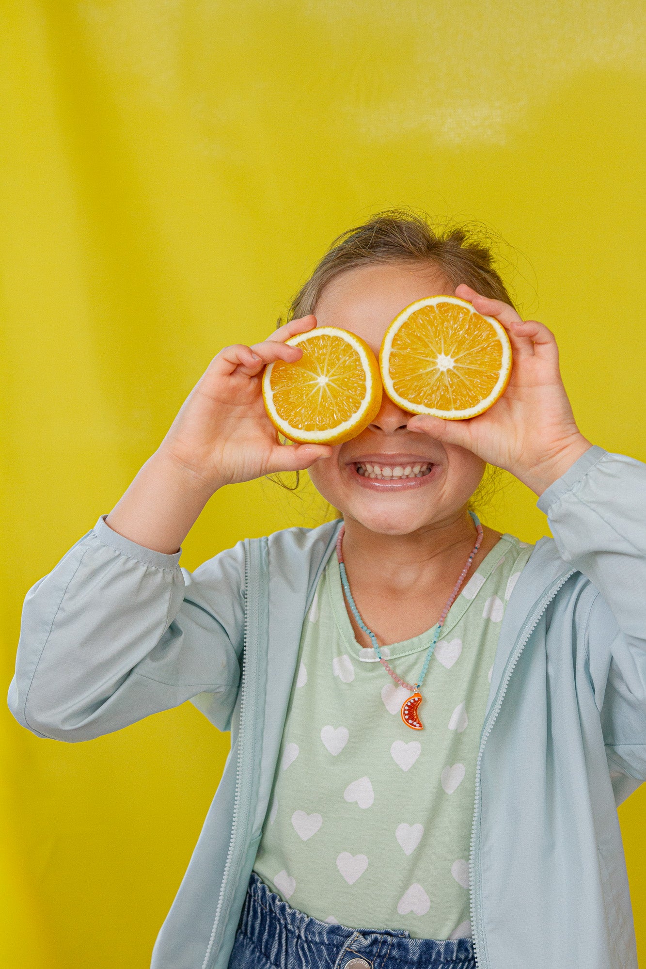 Niña con chaqueta de mezclilla y camiseta verde menta sostiene dos naranjas sobre los ojos frente a un fondo amarillo.