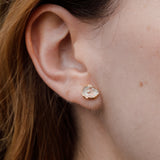 Primer plano lateral de la oreja de una mujer luciendo un arete Topos Cucu en bronce bañado en oro, pintado a mano en rosa con granulación de vidrio, cabello recogido detrás, sobre fondo neutro.