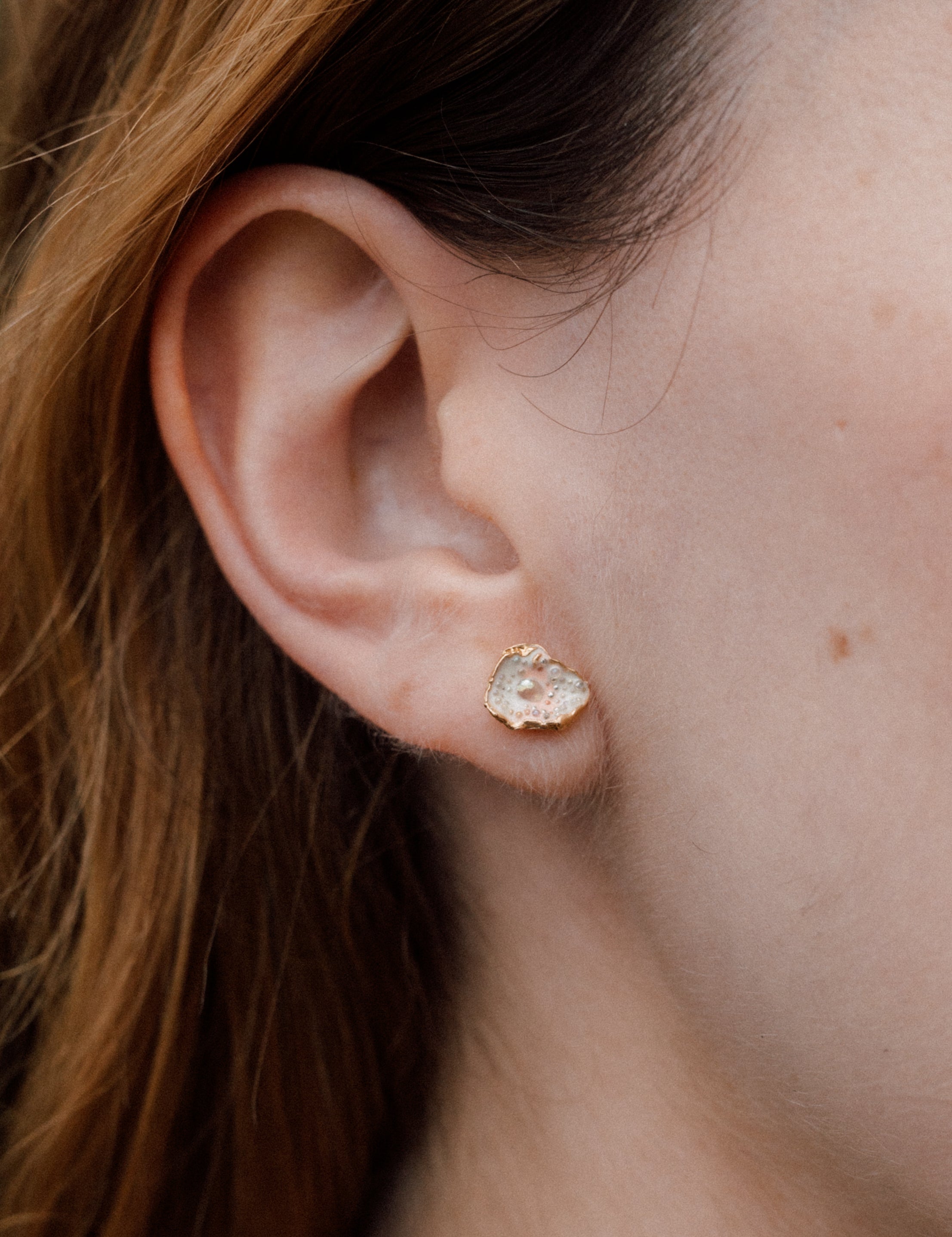 Primer plano lateral de la oreja de una mujer luciendo un arete Topos Cucu en bronce bañado en oro, pintado a mano en rosa con granulación de vidrio, cabello recogido detrás, sobre fondo neutro.