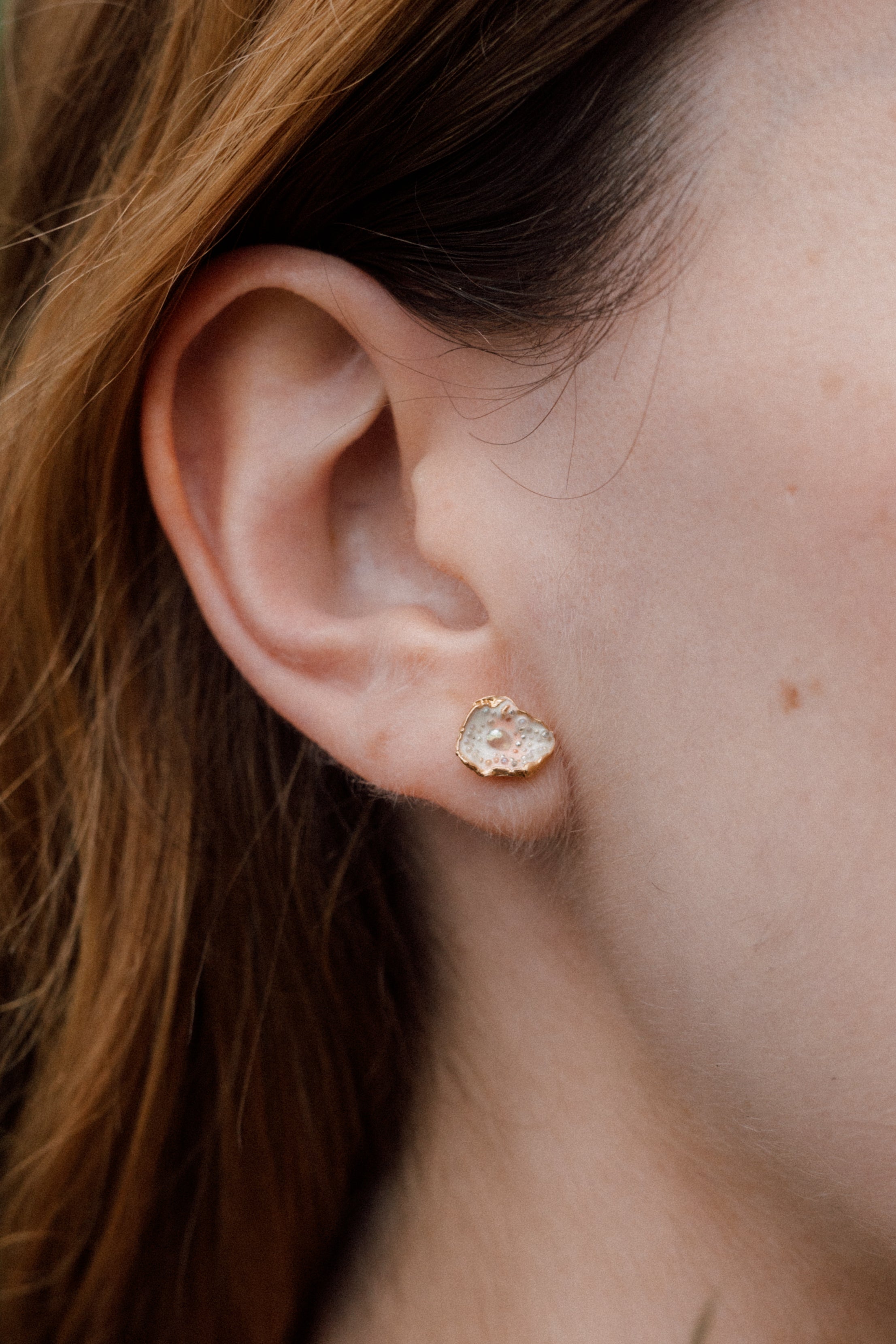 Primer plano lateral de la oreja de una mujer luciendo un arete Topos Cucu en bronce bañado en oro, pintado a mano en rosa con granulación de vidrio, cabello recogido detrás, sobre fondo neutro.