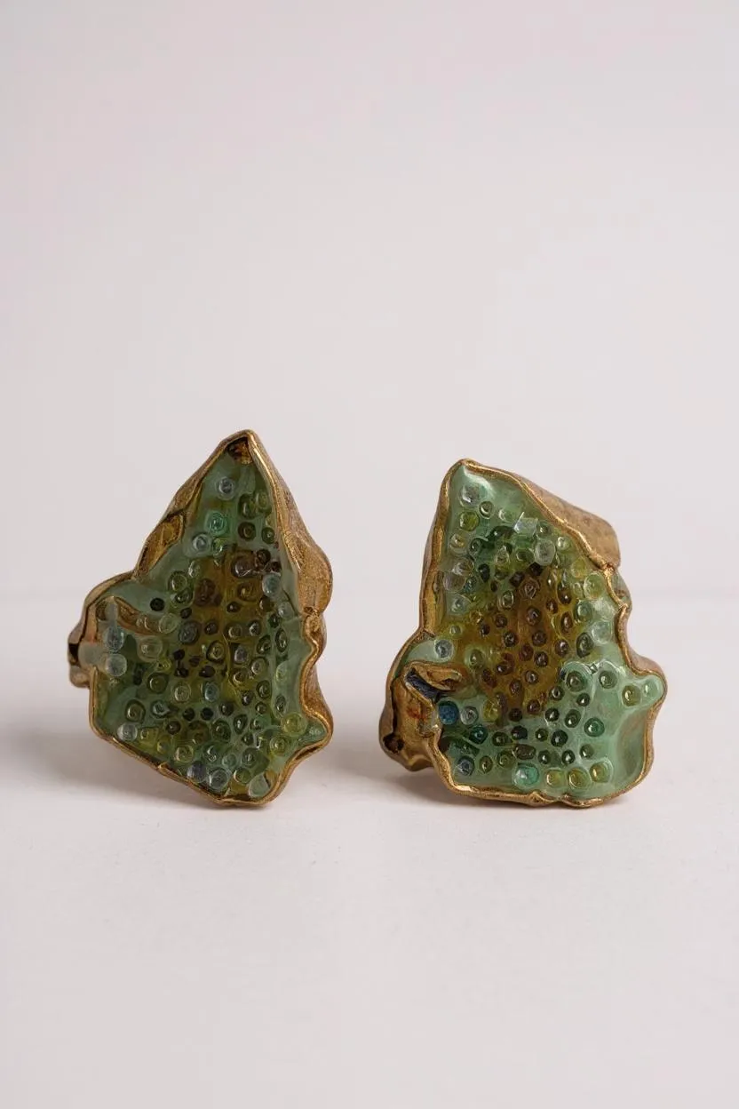 Disposición plana de dos aretes Topos Cucu en bronce bañado en oro, pintados a mano en verde salvia con granulación de vidrio, sobre fondo blanco.