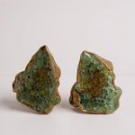 Disposición plana de dos aretes Topos Cucu en bronce bañado en oro, pintados a mano en verde salvia con granulación de vidrio, sobre fondo blanco.