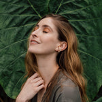 Retrato de una mujer con un arete Topos Cucu en bronce bañado en oro, pintado a mano en verde salvia con granulación de vidrio, posando frente a grandes hojas verdes.