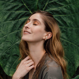 Retrato de una mujer con un arete Topos Cucu en bronce bañado en oro, pintado a mano en verde salvia con granulación de vidrio, posando frente a grandes hojas verdes.