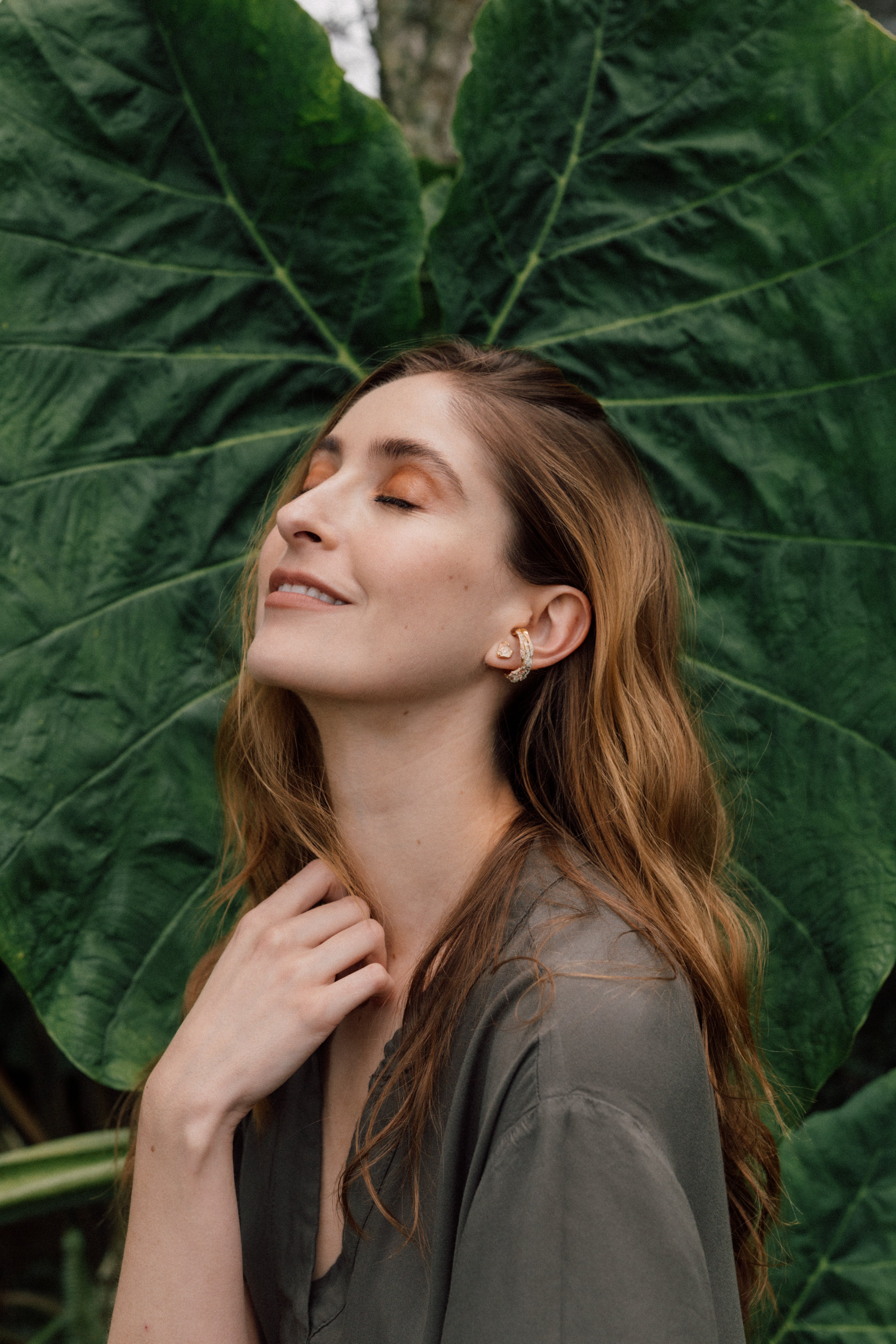 Retrato de una mujer con un arete Topos Cucu en bronce bañado en oro, pintado a mano en verde salvia con granulación de vidrio, posando frente a grandes hojas verdes.