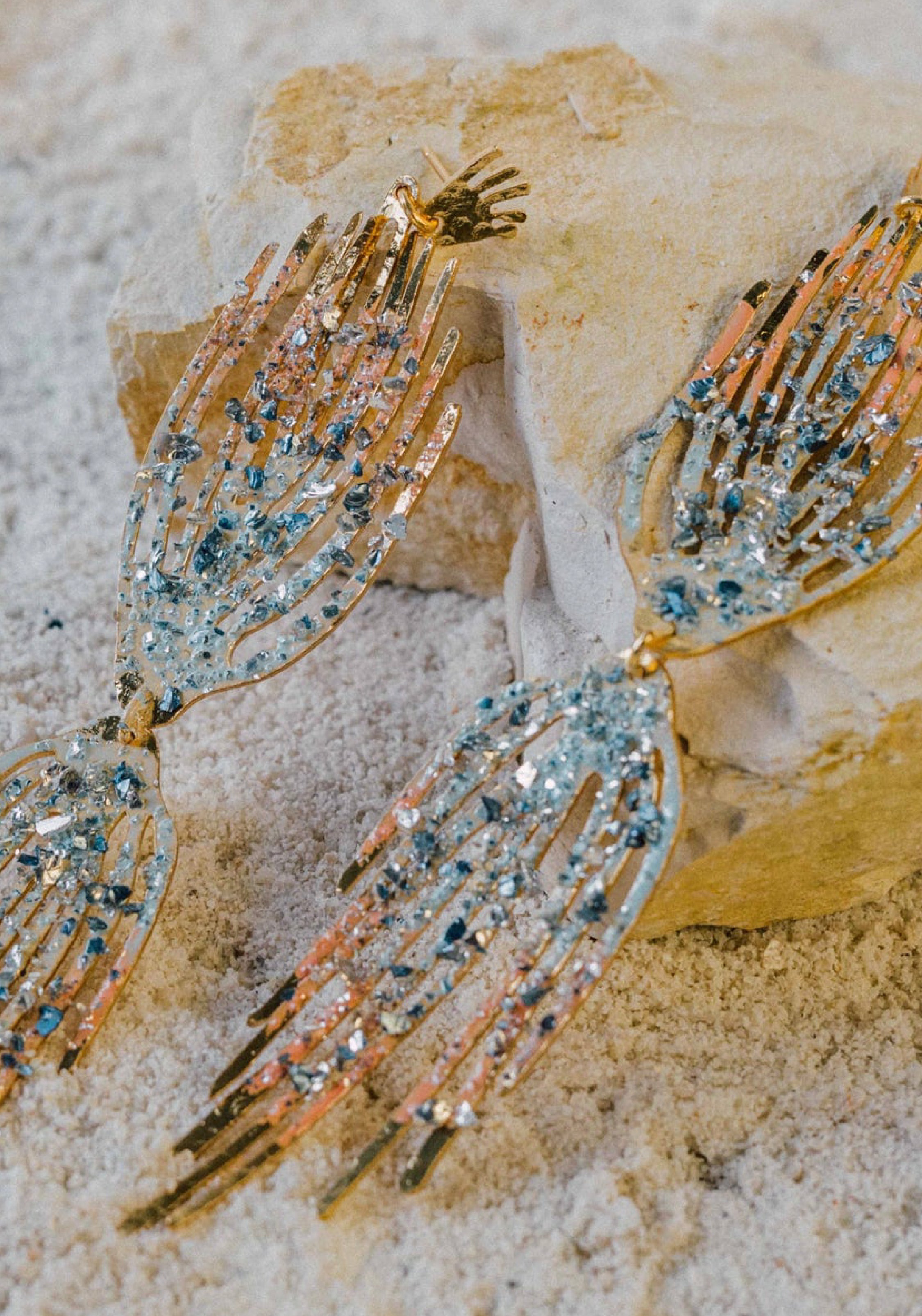 Par de pendientes artesanales de oro con forma de manos alargadas, adornados con cristales azules y rosas, expuestos sobre arena y piedra.