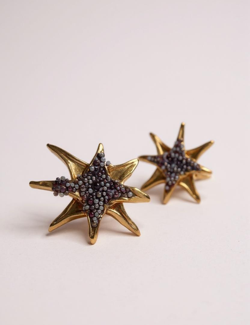 Par de aretes topos en forma de estrella bañados en oro con pequeños cristales púrpura, sobre fondo rosa pálido