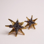 Par de aretes topos en forma de estrella bañados en oro con pequeños cristales púrpura, sobre fondo rosa pálido