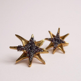 Par de aretes topos en forma de estrella bañados en oro con pequeños cristales púrpura, sobre fondo rosa pálido