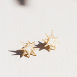 Dos aretes topos de estrella bañados en oro con detalles de cristales blancos, dispuestos sobre una superficie blanca con suaves sombras de hojas.