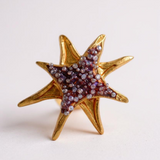 Un solo arete topos en forma de estrella bañados en oro con cristales púrpura, sobre fondo blanco limpio.