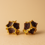 Primer plano de un par de aretes Galaxea en color rust: bañados en oro, con forma de estrella y cúmulos de gránulos de vidrio color óxido, sobre un fondo beige cálido.