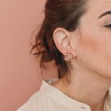  Perfil de una mujer luciendo un arete Galaxea en color Rust, bañado en oro con gránulos de vidrio, cabello recogido, sobre un fondo terracota suave.