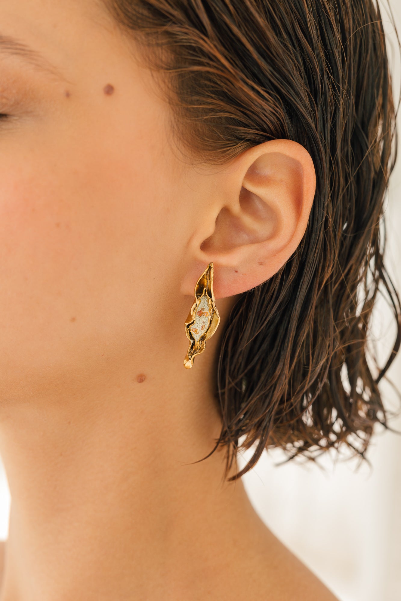 Primer plano lateral de la oreja de una mujer luciendo la variante Mostaza del Arete Lechuga, pintado a mano en amarillo mostaza con granulación de vidrio en montura de bronce bañado en oro, cabello recogido, sobre fondo neutro.