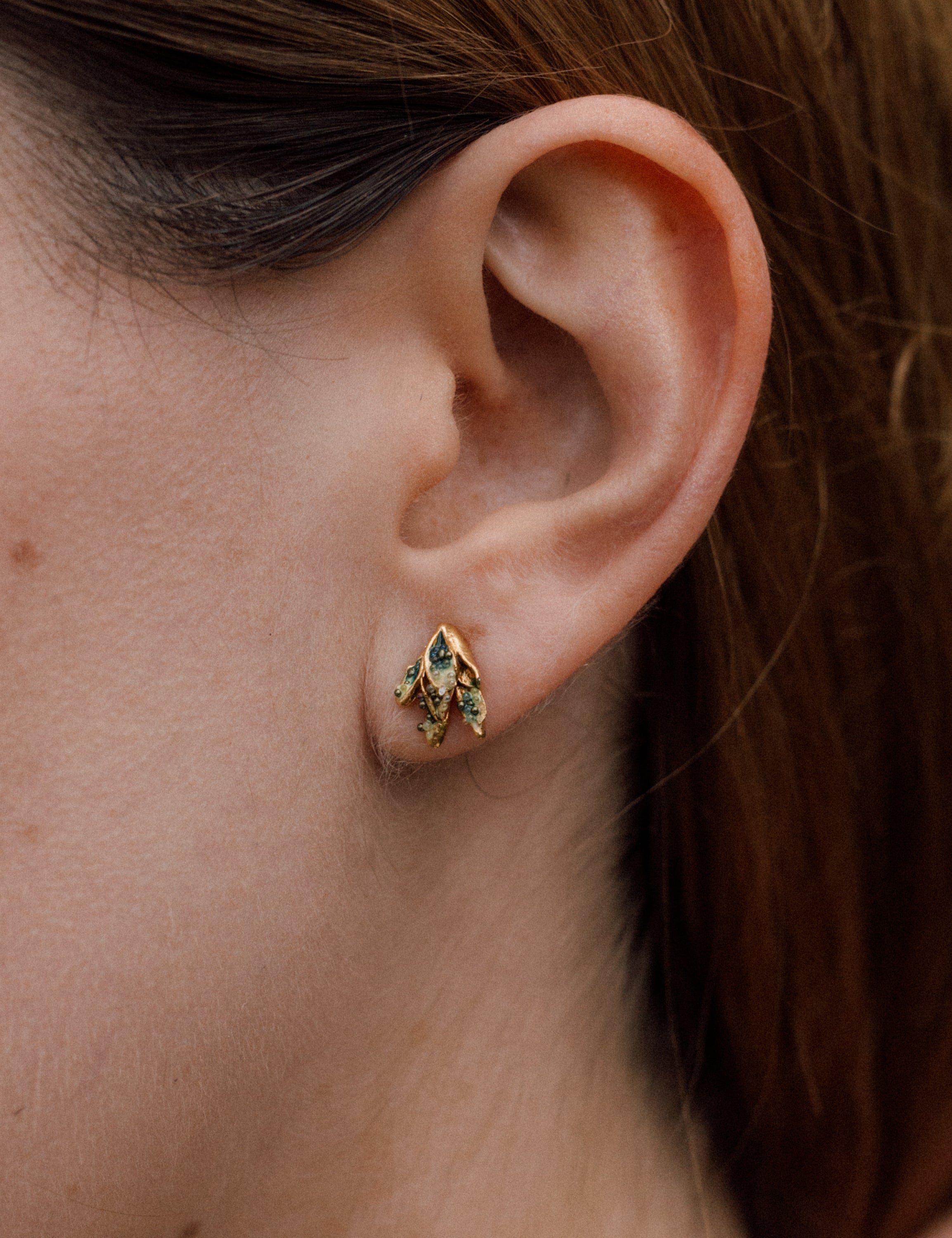 Primer plano lateral de la oreja de una mujer con arete topos Nebula Mini en bronce bañado en oro, pintado a mano en verde  con granulación de vidrio, cabello recogido contra un fondo neutro.