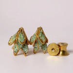 Disposición plana de dos aretes Topos Nebula Mini en forma de hoja pintados a mano en verde con granulación de vidrio, acompañados de un topo redondo de bronce bañado en oro colocado aparte, sobre superficie blanca.