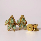 Disposición plana de dos aretes Topos Nebula Mini en forma de hoja pintados a mano en verde con granulación de vidrio, acompañados de un topo redondo de bronce bañado en oro colocado aparte, sobre superficie blanca.