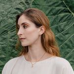 Retrato de una mujer con blusa blanca y un arete topos Nebula Mini en bronce bañado en oro, pintado a mano en verde con granulación de vidrio, frente a un fondo de grandes hojas verdes
