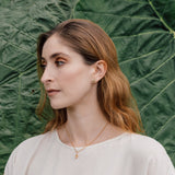 Retrato de una mujer con blusa blanca y un arete topos Nebula Mini en bronce bañado en oro, pintado a mano en verde con granulación de vidrio, frente a un fondo de grandes hojas verdes
