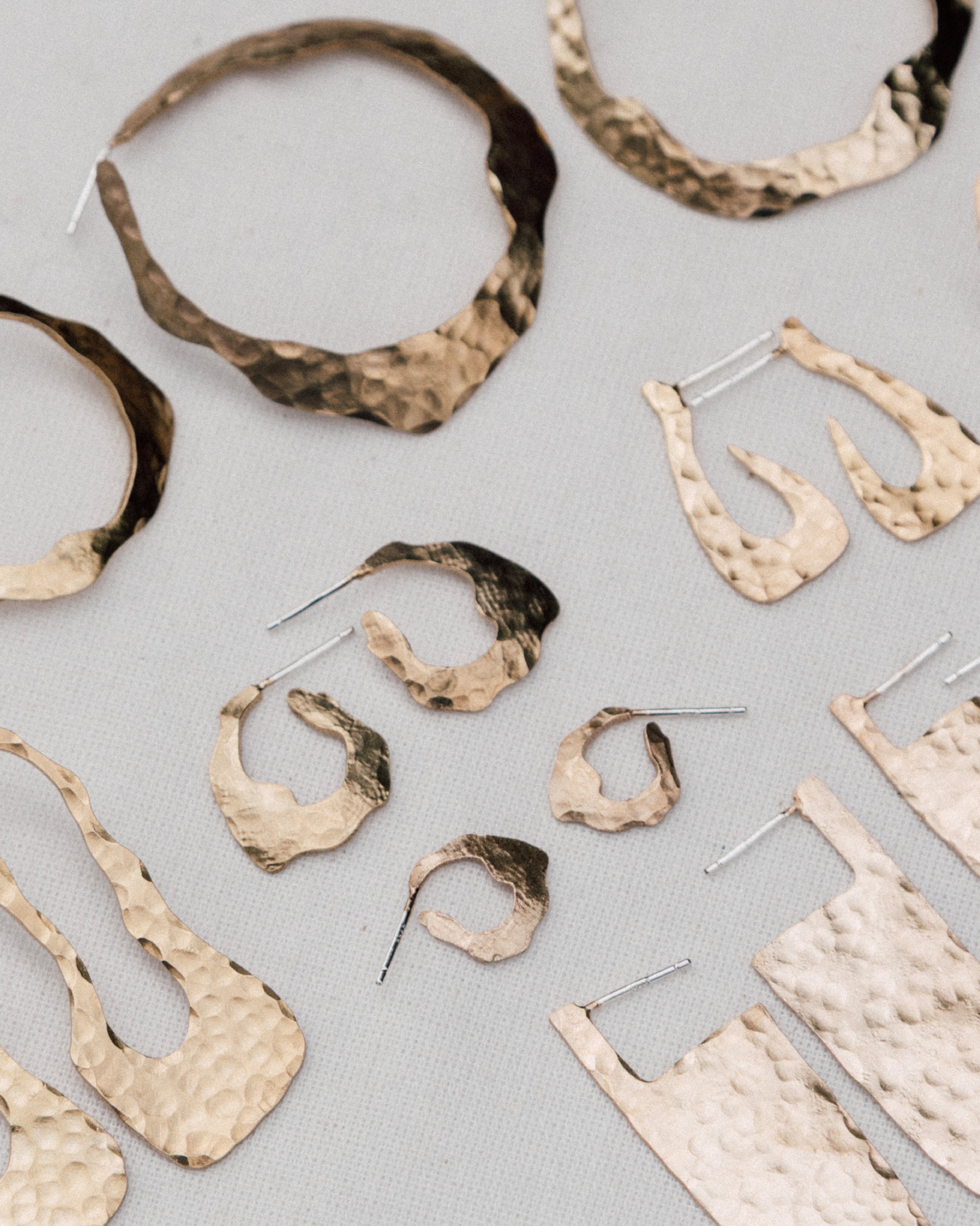 Surtido de pendientes artesanales de oro martillado con formas orgánicas, presentados sobre un fondo de tela neutra.