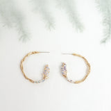 vista lateral de Aros Candonga Espiga en bronce bañado en oro con detalles lavanda pintados y granulación de vidrio, sobre fondo botánico difuminado.