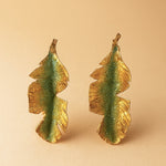 Par de aretes Hojas de Plátano en bronce bañado en oro pintado a mano en verde sage detalle de granulación de vidrio, sobre fondo durazno suave.