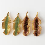 Disposición plana de cuatro aretes Hojas de Plátano alineados horizontalmente sobre fondo blanco—dos pintados a mano en verde salvia y dos en rust—con granulación de vidrio y montura de bronce bañado en oro.